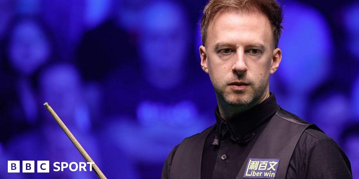 Judd Trump in black shirt and waistcoat carrying a snooker cue