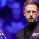 Judd Trump in black shirt and waistcoat carrying a snooker cue